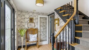 A staircase with a wooden handrail and black balusters, a wooden bench with a cushion, a wall with floral wallpaper, and a door with a mirror.