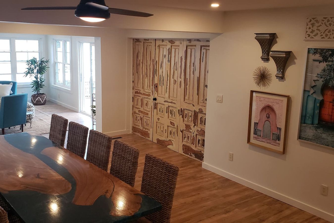 A dining area with a large wooden table and wicker chairs, a ceiling fan, and a door with a unique textured design.