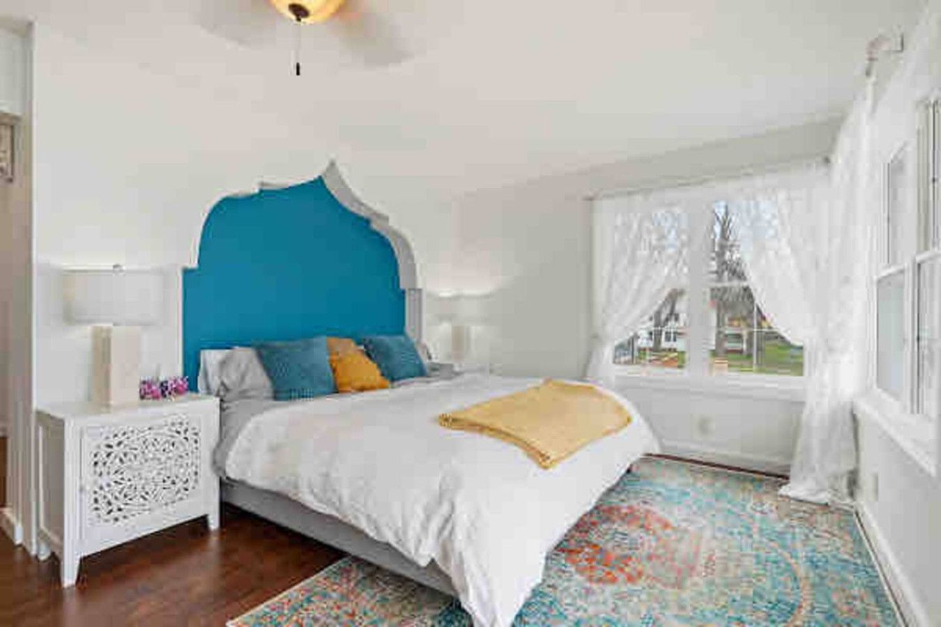 A bedroom with a bed, a nightstand, a window with curtains, and a view of the outside.