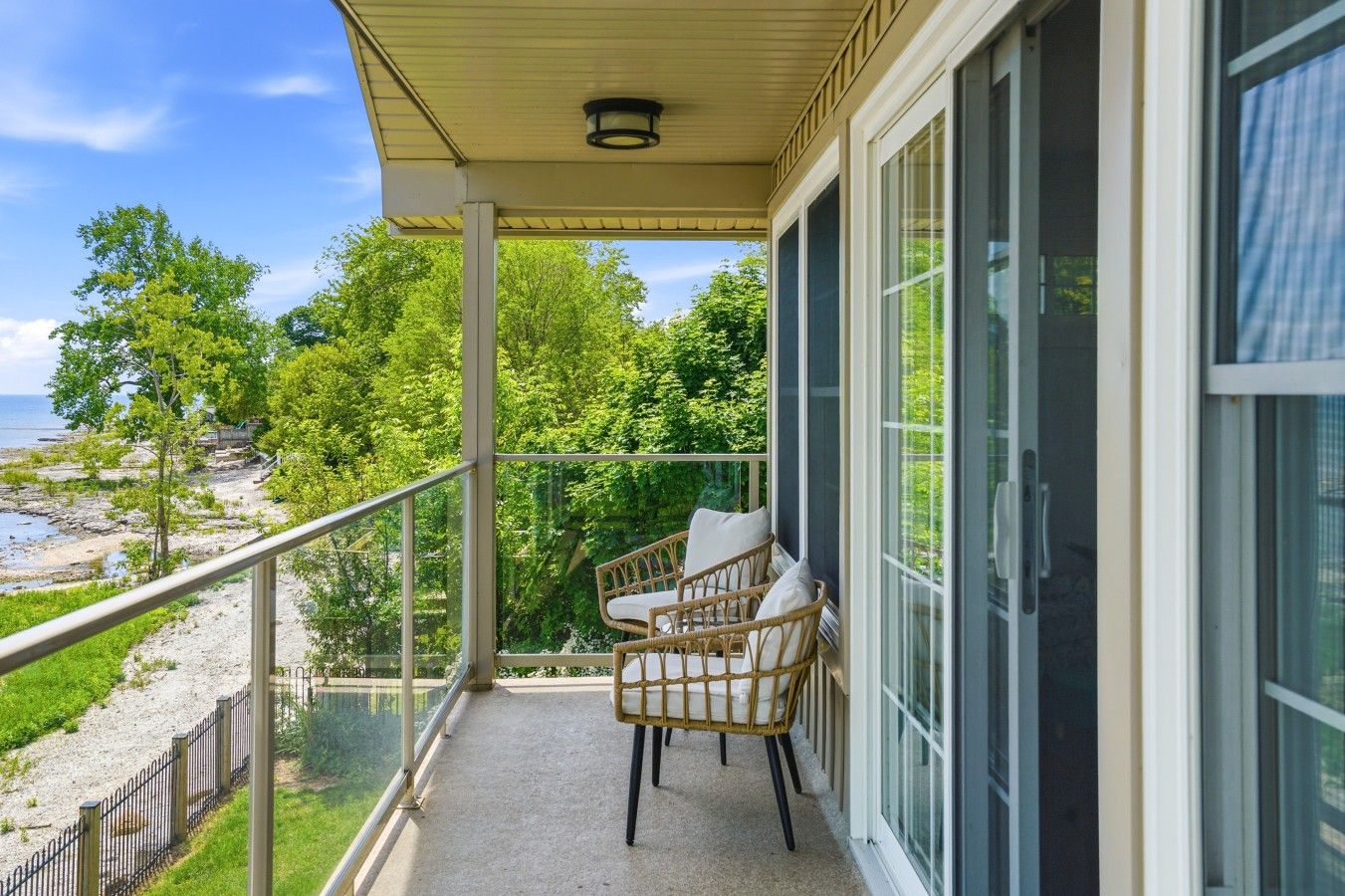 A balcony with a view of a beach and trees, featuring two wicker chairs and a glass railing.