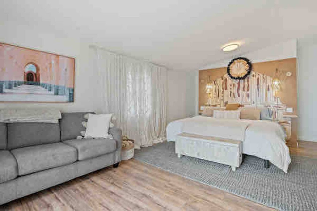 A bedroom with a large bed, a grey sofa, and a painting on the wall.