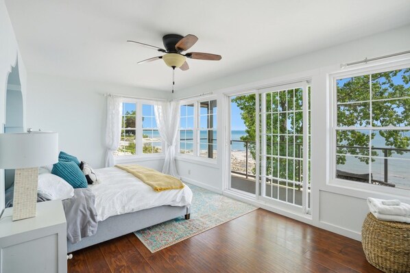 A bedroom with a bed, a ceiling fan, a window with a view of the beach, and a wicker ottoman.
