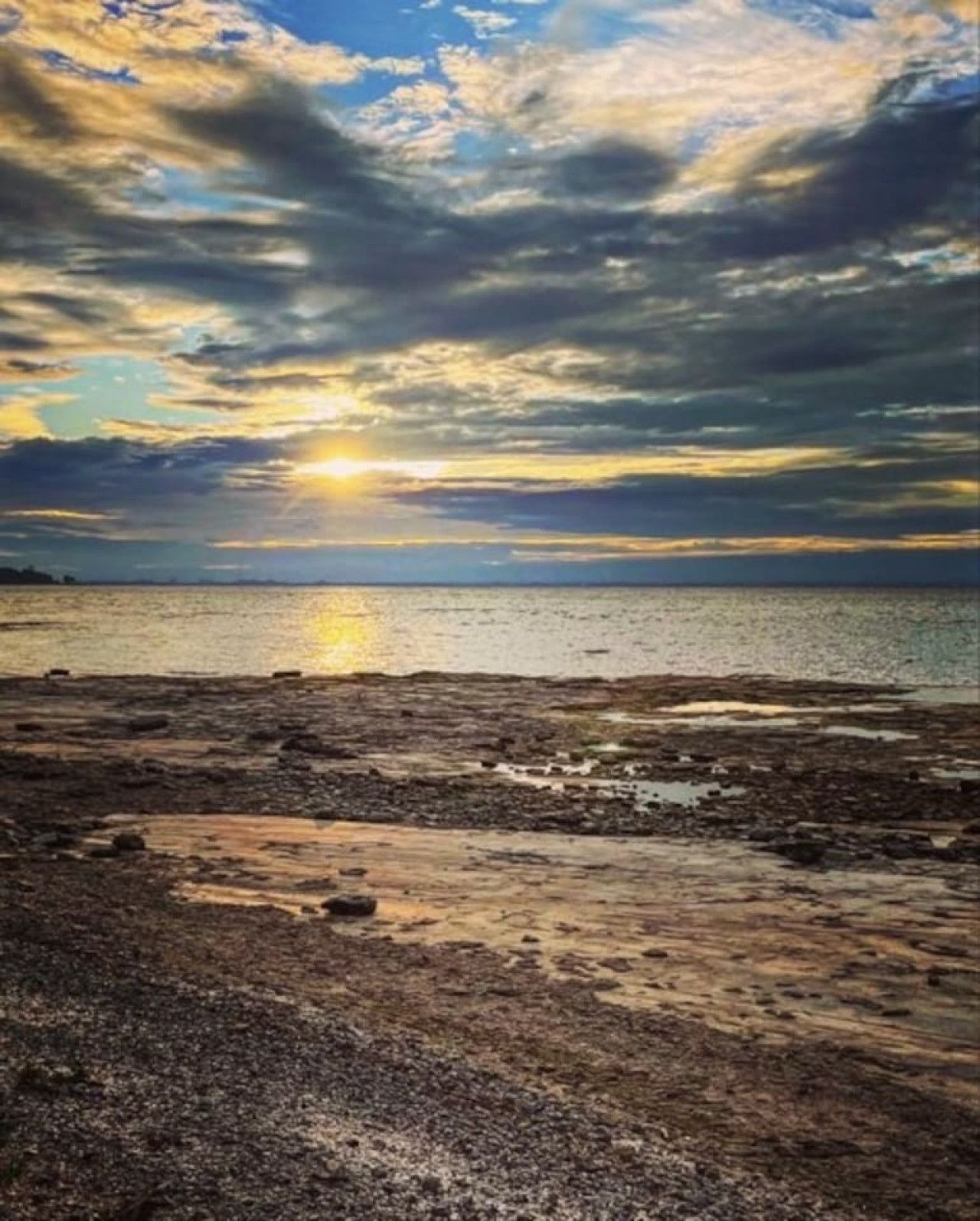 A beach with a sunset over the water, rocky foreground, and a clear sky.