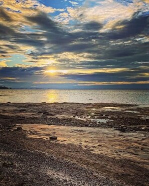 A beach with a sunset over the water, rocky foreground, and a clear sky.