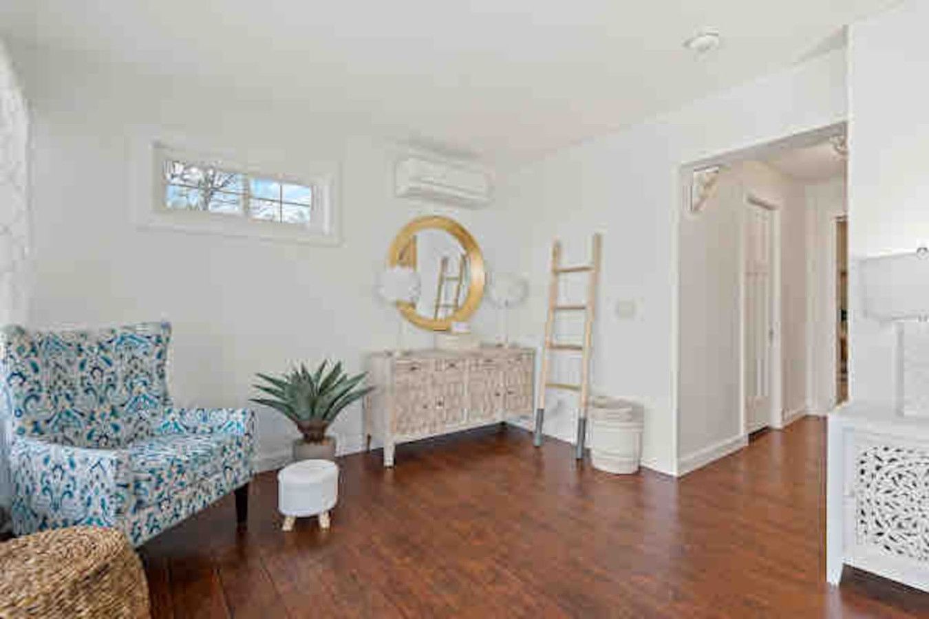 Una habitación amplia con piso de madera, un sillón estampado, un pequeño reposapiés blanco, un espejo decorativo y un mueble blanco.