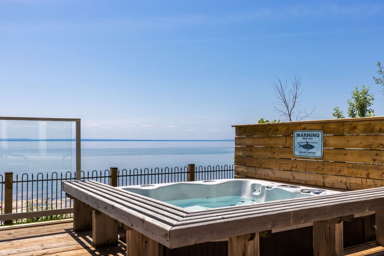 A wooden deck with a hot tub overlooking a body of water, a warning sign about shark sightings, and a clear sky.