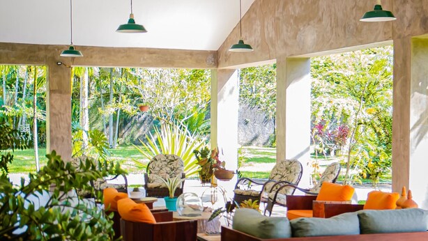 A spacious living room with a wooden floor, a sofa with cushions, and a coffee table. There are hanging green pendant lights and large windows with a view of greenery outside. - Casa c Wifi Churrasq Pisc red rio Pet Friendly (Benevides)