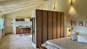 A bedroom with a bed, a bench with towels, a wooden wardrobe, and a kitchen area with stools and a counter. - Haven of peace in the Garraf: 18th-century masia, sea view & exceptional light (Olivella)