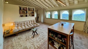 A spacious room with a high ceiling, a built-in bookshelf, a cushioned seating area, and large windows offering a view of the outdoors. - Haven of peace in the Garraf: 18th-century masia, sea view & exceptional light (Olivella)