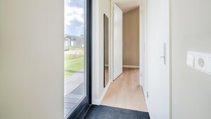 Un couloir avec une porte vitrée donnant sur un jardin, un plancher en bois et un mur blanc avec un interrupteur.