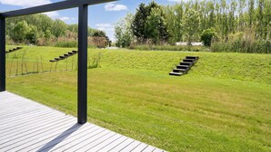 Une terrasse en bois avec une rambarde en métal, donnant sur une pente herbeuse où descendent des marches.