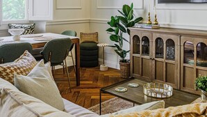 Een woonkamer met een bank, een salontafel, een eethoek met stoelen, een aan de muur bevestigde televisie en een groot raam met uitzicht op groen.