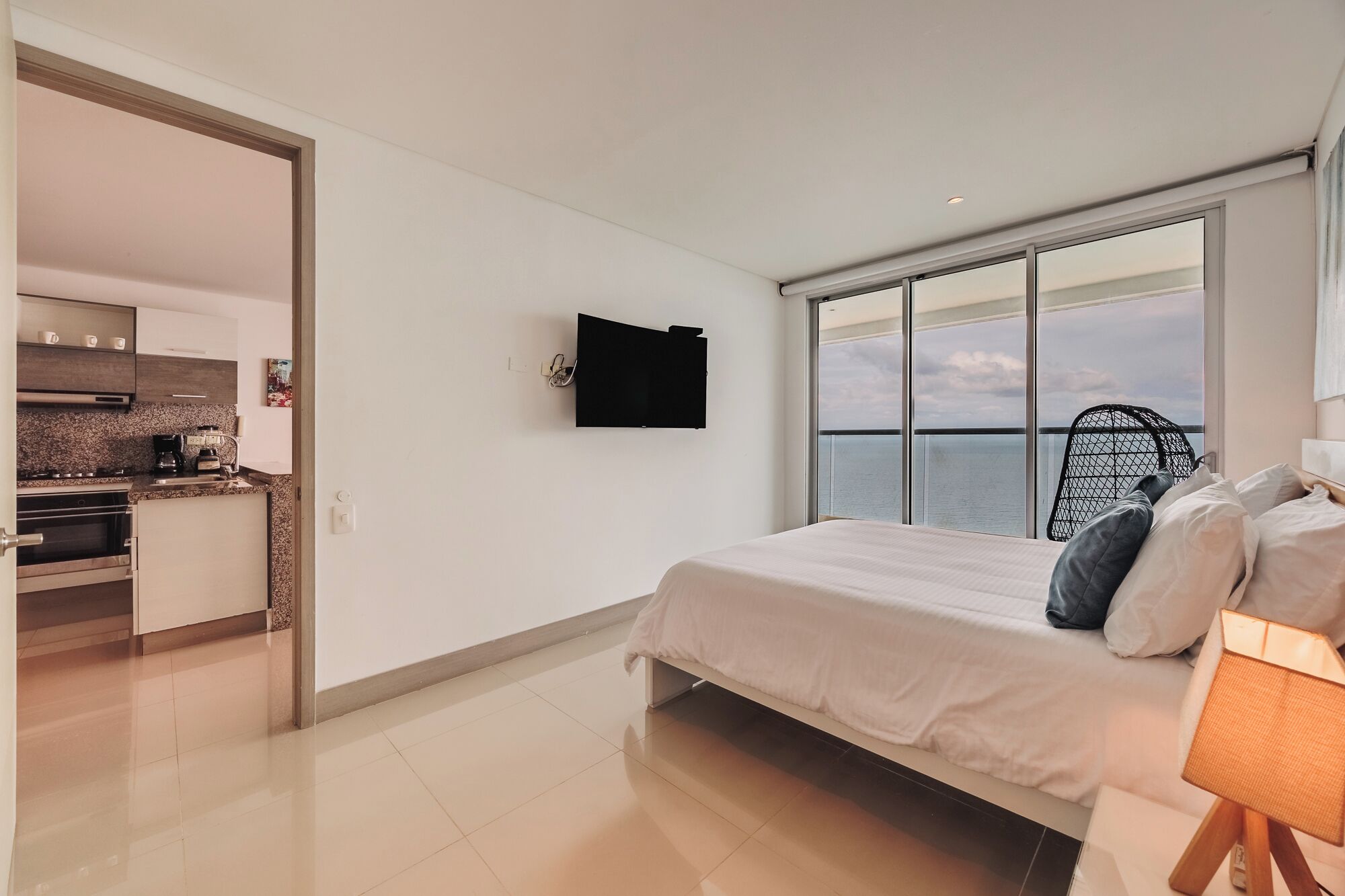 A modern bedroom with a large window, a bed, and a kitchenette in the adjacent room.