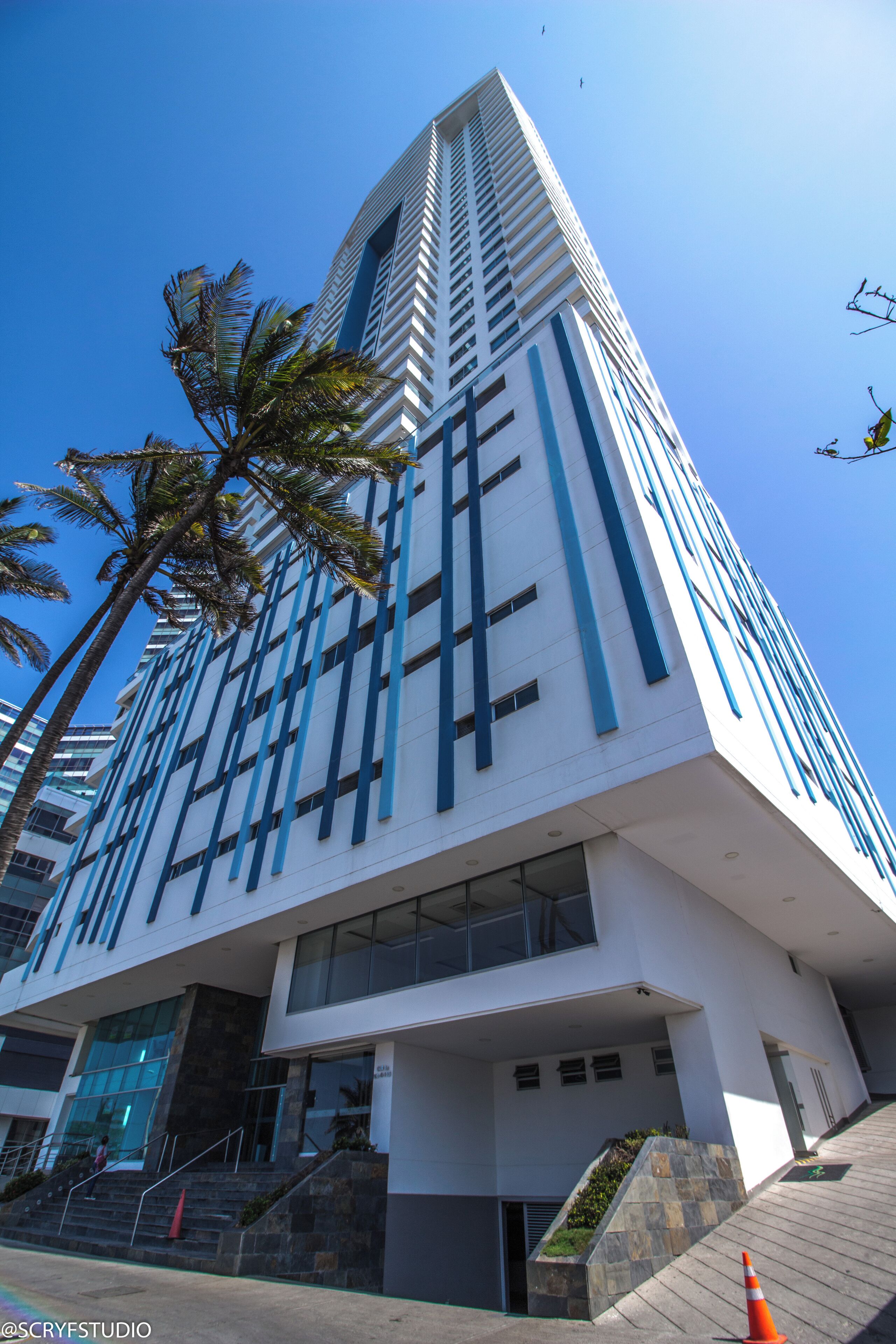 A modern building with a white facade and blue accents, featuring a staircase and palm trees.