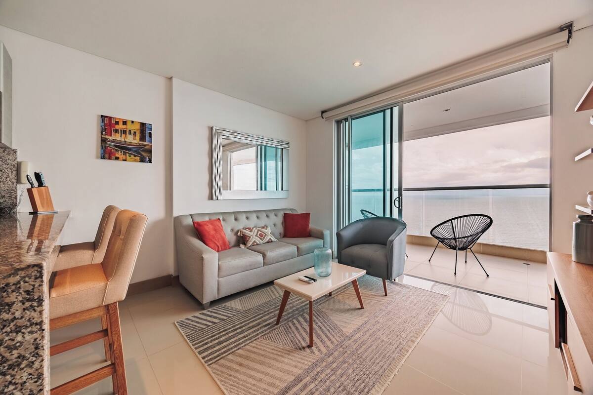A modern living room with a grey sofa, a coffee table, and a chair. There is a large window with a view of the sea.