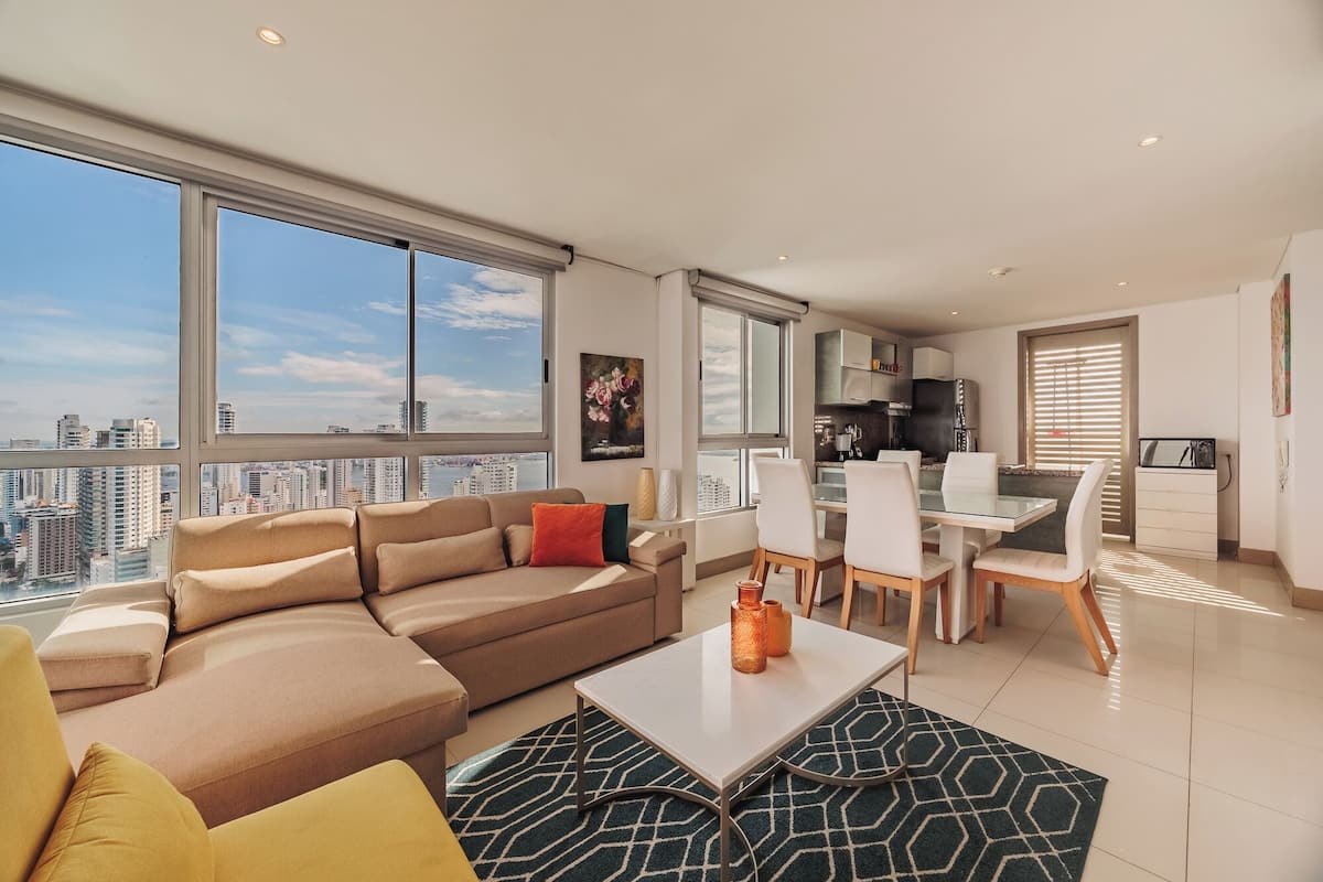 A modern living room with a sofa, a dining table, and chairs. There is a kitchen area with a refrigerator and cabinets. Large windows offer a view of the city.