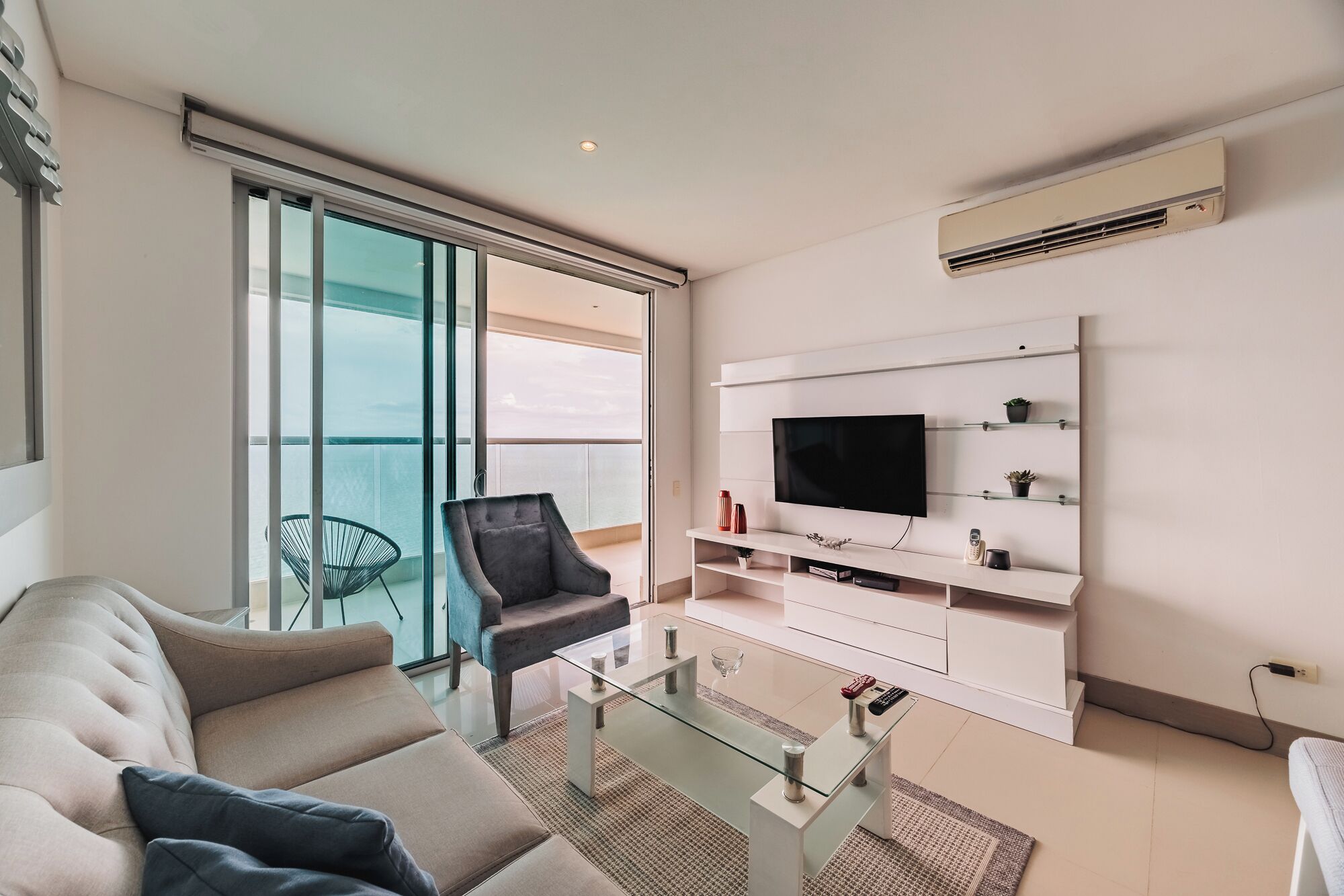 A modern living room with a sofa, a glass coffee table, a TV mounted on the wall, and a balcony with a chair.