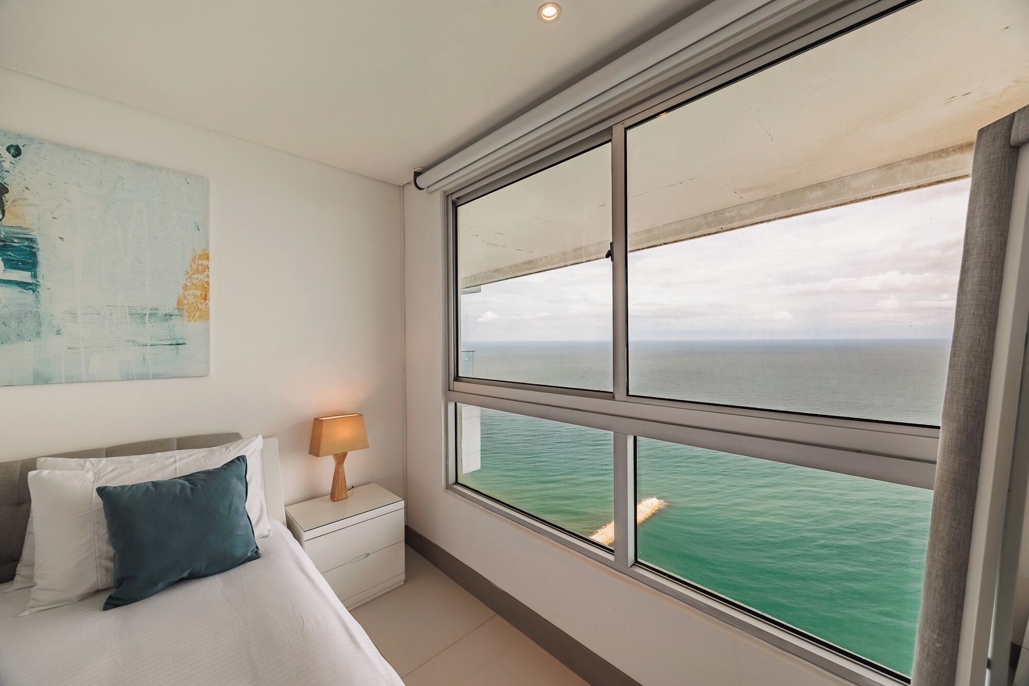 Une chambre avec une grande fenêtre donnant sur la mer, une lampe de chevet et un lit avec des oreillers.