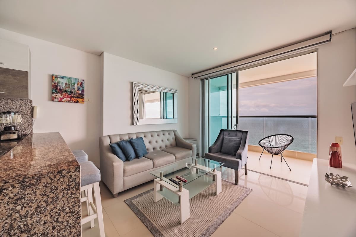 A modern living room with a sofa, coffee table, and chairs, a kitchen area with a blender, and a balcony with a view of the sea.