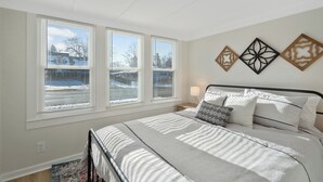 A bedroom with a bed, white walls, three windows with blinds, a nightstand with a lamp, and decorative wall art. - Charming 3-bedroom house in North Syracuse (North Syracuse)