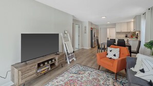 A modern living room with a flat-screen TV, a wooden entertainment unit, an orange armchair, a gray sofa, and a colorful rug. - Charming 3-bedroom house in North Syracuse (North Syracuse)