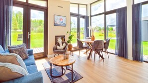Een ruime woonkamer met een blauwe bank, een houten salontafel en een eethoek met stoelen en een tafel. Grote ramen bieden uitzicht op de omgeving.