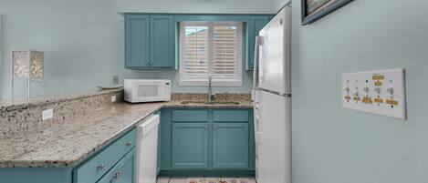 A kitchen with teal cabinets, a white microwave, a refrigerator, and a window with blinds.