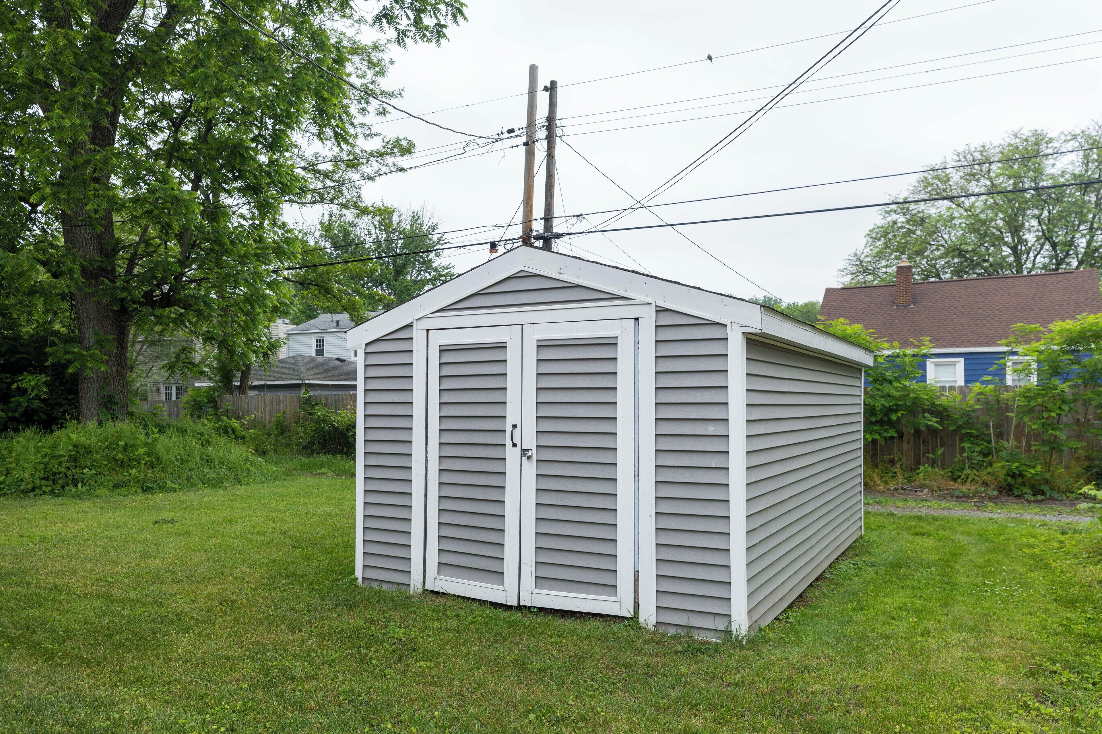 A small, single-story shed with a gabled roof and a door, surrounded by a grassy yard.