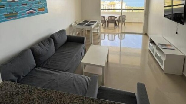 Un saló modern amb un sofà gris, una taula de centre i un televisor a la paret. Hi ha un quadre amb vaixells a la paret i un balcó amb vista al mar.