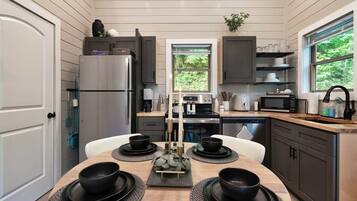 A modern kitchen with a dining area, featuring a stainless steel refrigerator, microwave, stove, and a window providing a view of greenery.
