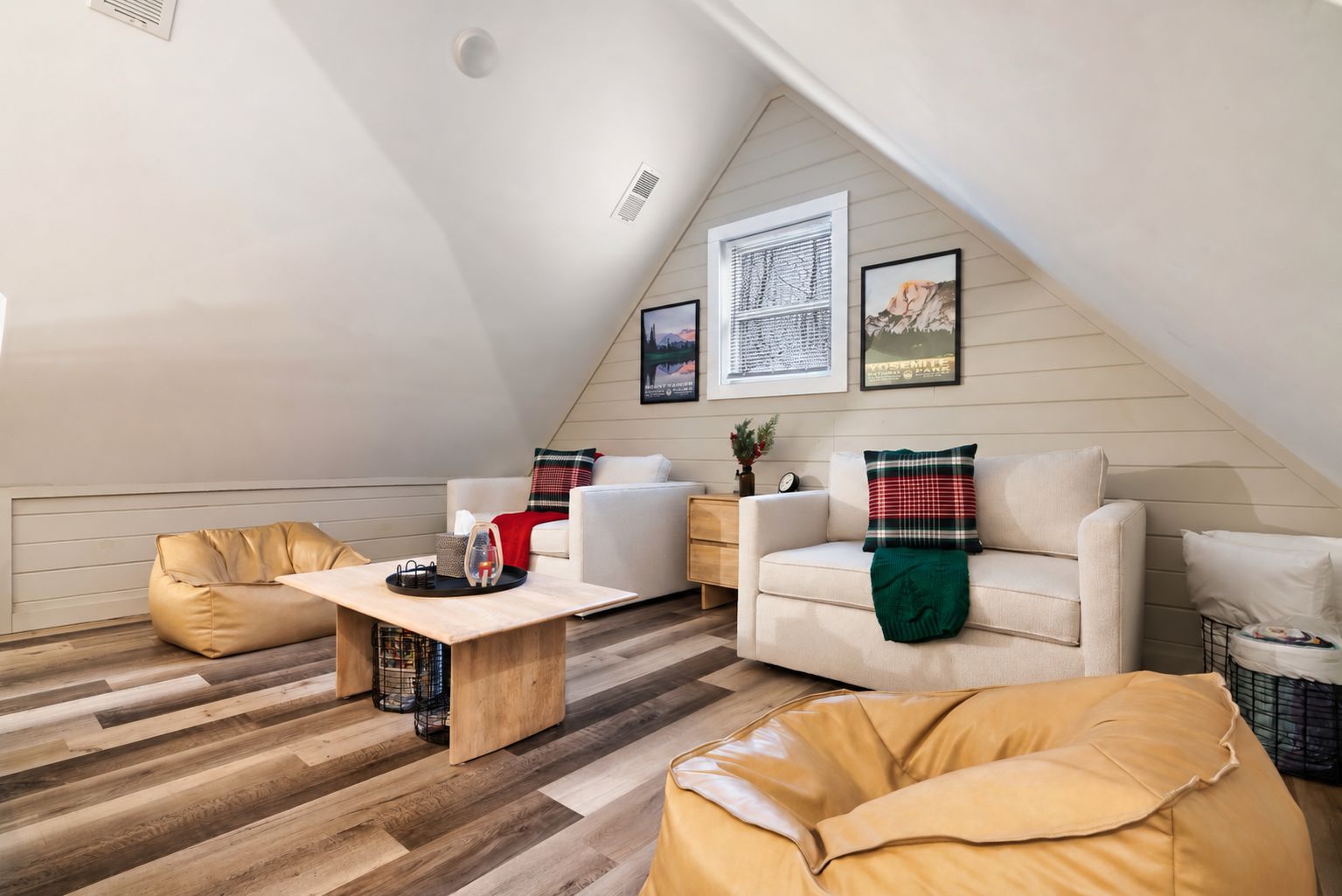 Un salón estilo loft con techo inclinado, piso de madera y una variedad de asientos, incluyendo un sofá y pufs.