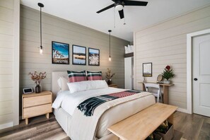 A bedroom with a bed, bedside tables, a bench, a ceiling fan, wall-mounted lights, and framed pictures.