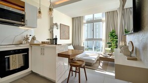 A modern kitchen with a built-in oven, a dining area with a wooden table and stools, a bed with a view of the city, and a large window with curtains. - Caribbean Coastal View Apartment (Playa Juan Dolio)
