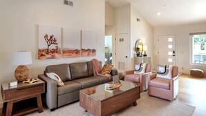 A spacious living room with a brown leather sofa, a glass coffee table, and a pink armchair. There are three framed artworks on the wall, a lamp on a side table, and a large window providing natural light.