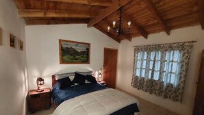 A bedroom with a bed, wooden ceiling beams, a painting, a nightstand, and a window with curtains.