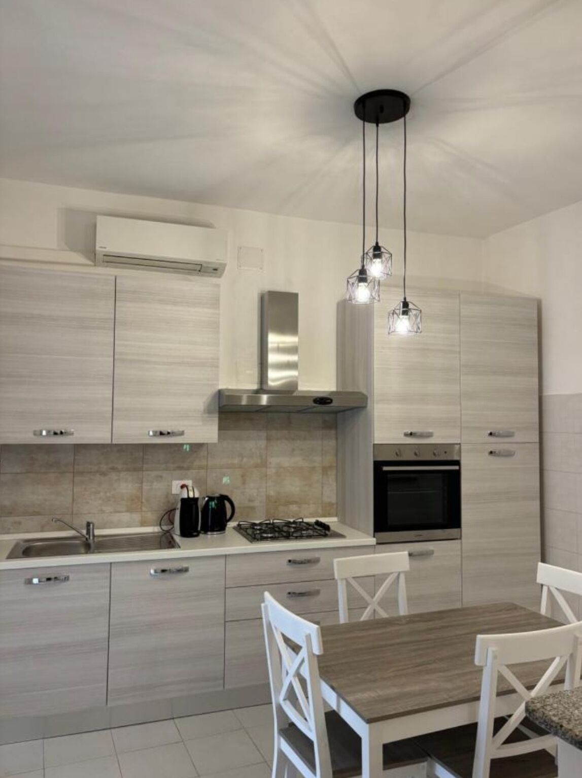 A modern kitchen with light wood cabinetry, a stainless steel range hood, a built-in oven, and a dining area with a wooden table and white chairs.