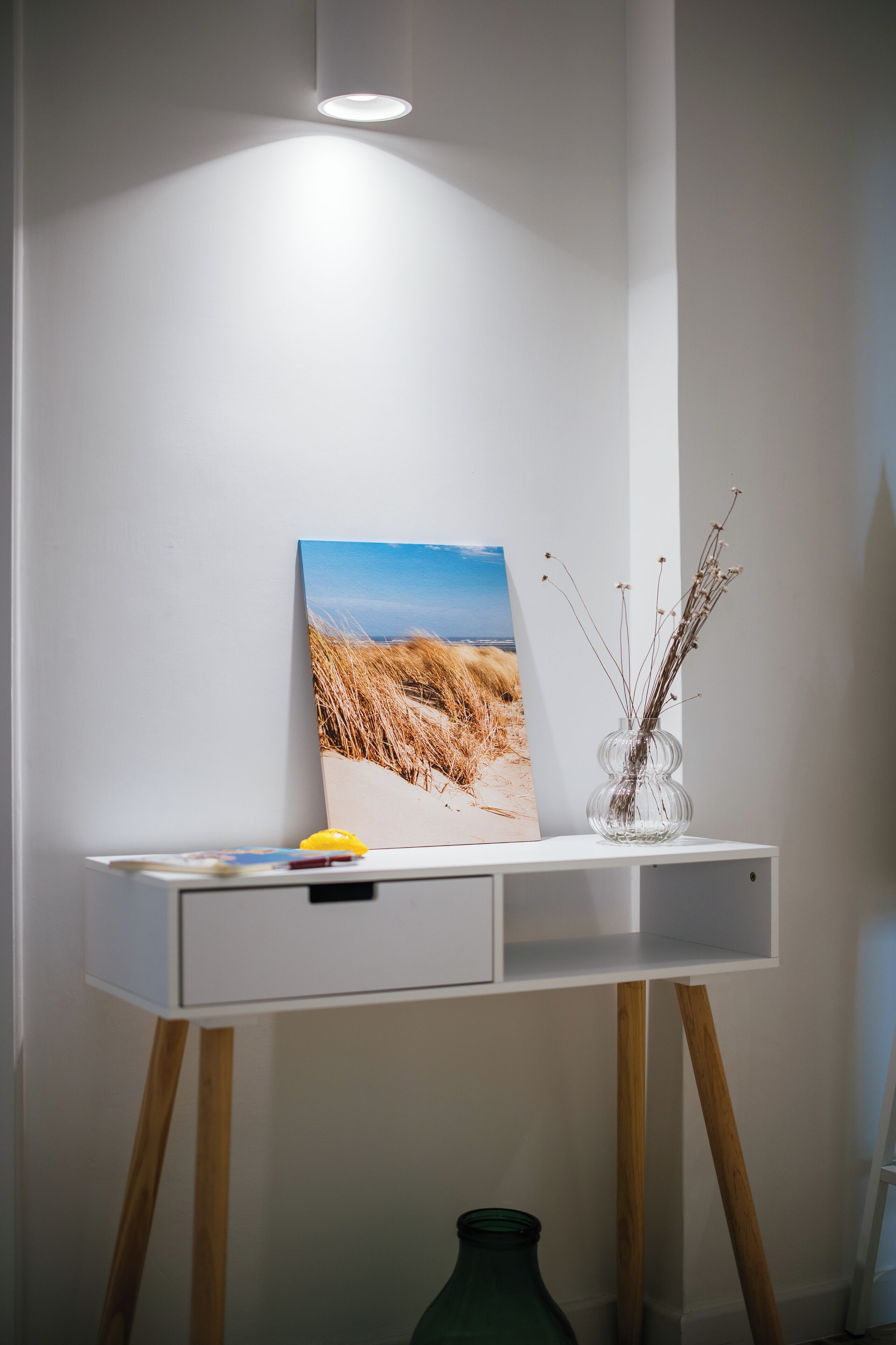 A white desk with a painting of a beach scene, a clear glass vase with dried plants, and a green bottle.