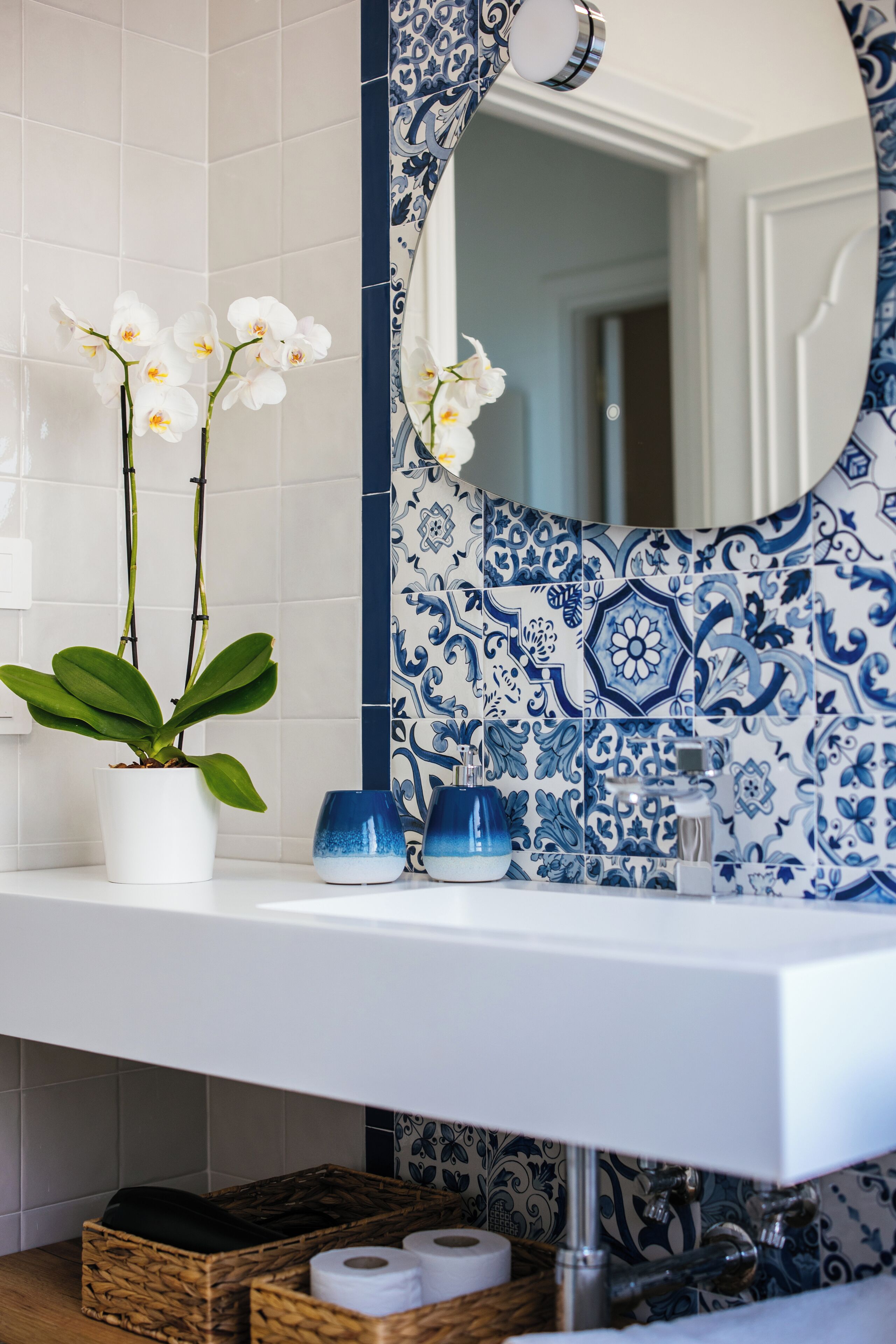 Ein Badezimmer mit einem weißen Waschbecken, blau-weißen Fliesen mit Muster, einem Spiegel mit dekorativem Rahmen und einem Orchideenstrauch im Topf.