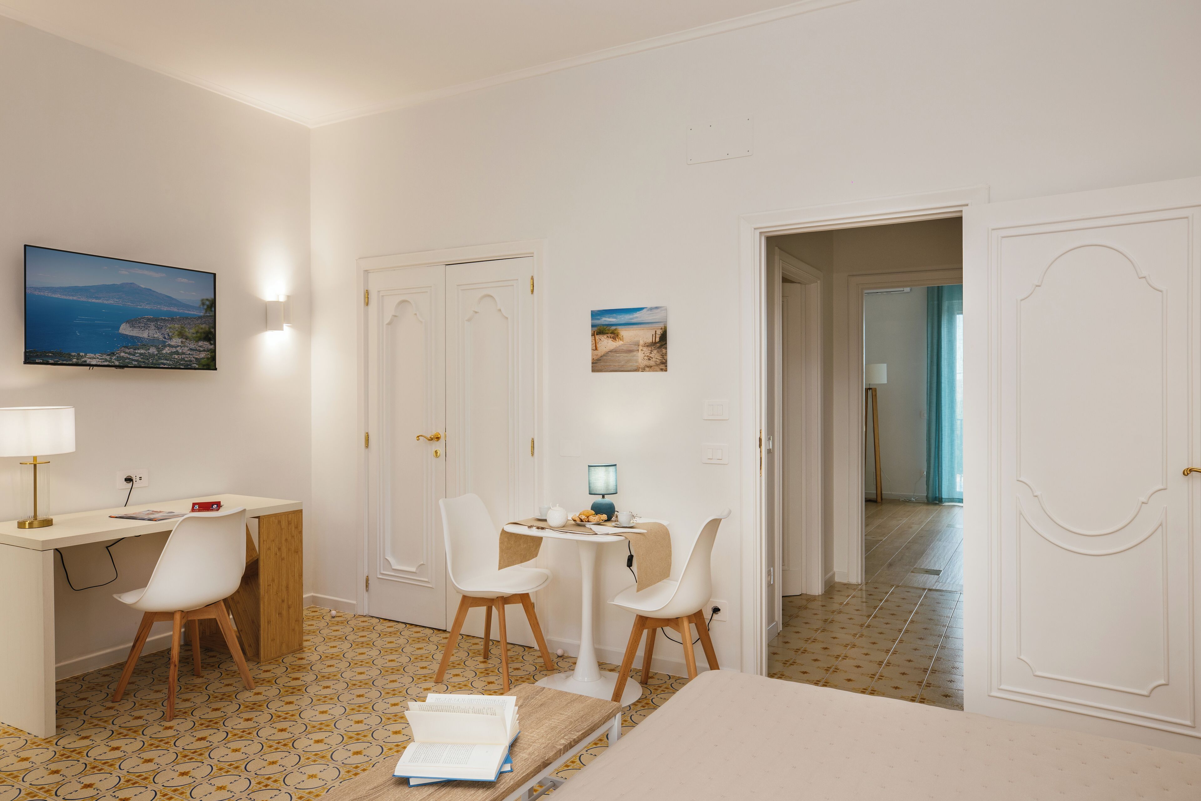 A room with a desk, two chairs, a bed, and a small table with a lamp. There are two doors, one leading to another room and another to the outside.
