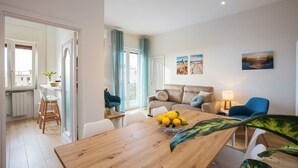 A modern living room with a wooden dining table, a sofa, and a blue armchair. There is a window with curtains, a lamp, and a painting on the wall. - De Vivo Realty - Lunamare Sorrento (Sorrento)