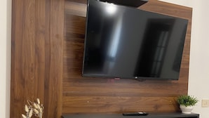 A modern living room with a flat-screen TV mounted on a wooden wall, a black entertainment unit below, and two potted plants. - Vacation Home Balai Porto Torres (Porto Torres)