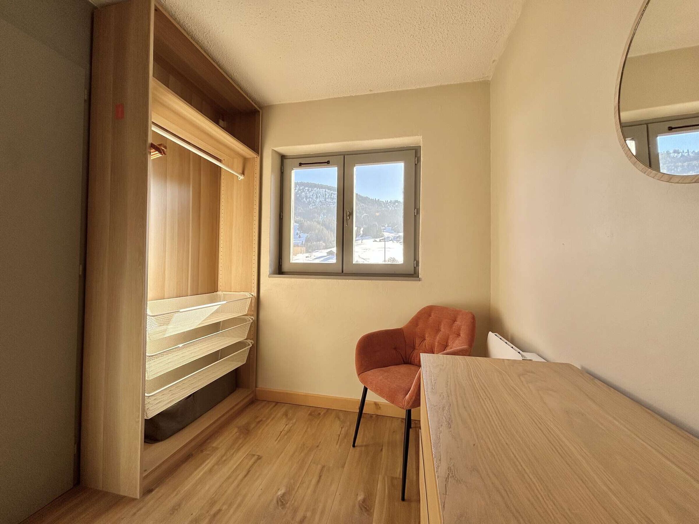 Una habitación pequeña con un armario de madera, una ventana con vistas a un paisaje nevado, una sola silla naranja y una mesa de madera.