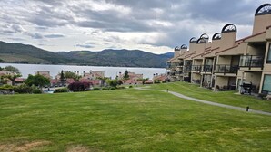Zona verde con vistas a un lago y montañas al fondo. Se observan edificios con balcones y una piscina.