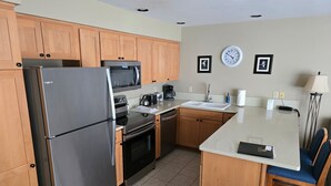 Una cocina compacta con armarios de madera, un refrigerador de acero inoxidable, un microondas, un horno, un lavamanos y un pequeño comedor con una silla.