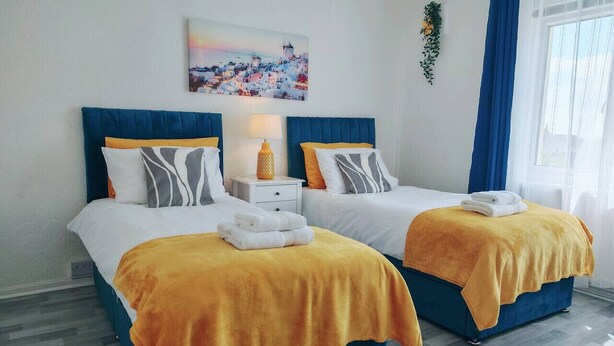 A hotel room with two beds, each with a blue headboard and white bedding. There is a painting on the wall above the beds, a small table with a lamp, and a window with blue curtains. - Family and pet-friendly flat near beach and countryside (Baglan)