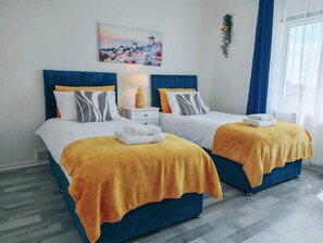 Habitación de hotel con dos camas, cada una con cabecero azul y ropa de cama blanca. Hay un cuadro en la pared sobre las camas, una mesita con una lámpara y una ventana con cortinas azules.