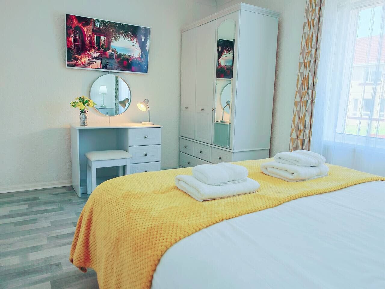 A hotel room with a bed, a bedside table with a lamp, a mirror, a wardrobe, and a painting on the wall.