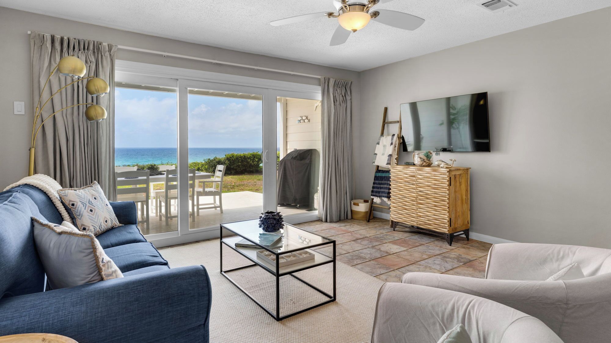 Ein Wohnzimmer mit einer Couch, einem Couchtisch und einem Fernseher. Eine Glastür führt auf einen Balkon mit Blick aufs Meer.