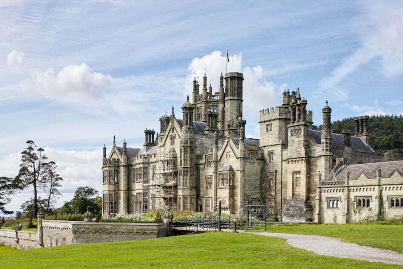 A large, historic stone building with multiple towers and turrets, surrounded by a well-maintained lawn and a small bridge.