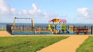 Un parque infantil con tobogán y columpios, rodeado por una valla de metal, con un camino que lleva a él.